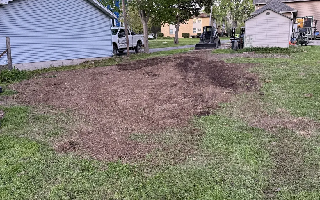 Levelled bare soil beside a garage where a stump was ground out, ready to seed